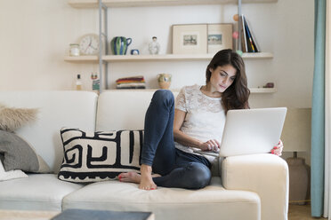 Woman sitting on couch at home using laptop - KKAF00385