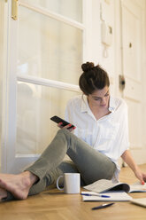 Junge Frau sitzt mit Kaffeetasse und Smartphone auf dem Boden und schaut in eine Broschüre - KKAF00391