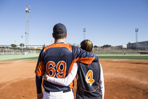 Rückansicht eines männlichen und eines weiblichen Baseballspielers, die sich auf einem Baseballfeld umarmen, lizenzfreies Stockfoto