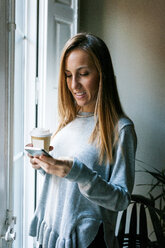 Junge Frau am Fenster mit Mobiltelefon und Kaffee zum Mitnehmen - VABF01099