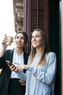 Two young women on balcony with cell phone and takeaway coffee looking up - VABF01101