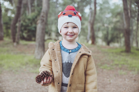 Porträt eines Jungen mit Wollmütze, der Tannenzapfen im Wald hält, lizenzfreies Stockfoto