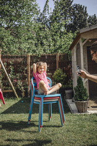 Father taking cell phone picture of daughter sitting on chair in garden - JOSF00578