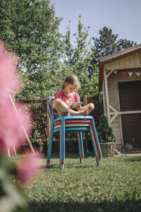 Mädchen sitzt auf einem Stapel von Stühlen im Garten und benutzt ein Tablet - JOSF00580