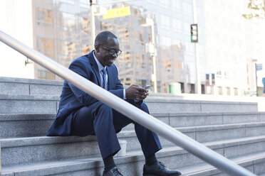 Lächelnder Geschäftsmann, der auf einer Treppe sitzt und sein Mobiltelefon überprüft - WESTF22654