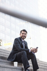 Smiling businessman sitting on stairs with cell phone and takeaway coffee - WESTF22662