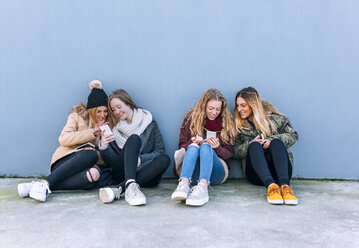 Four friends sitting side by side on the ground looking at cell phones - MGOF02923