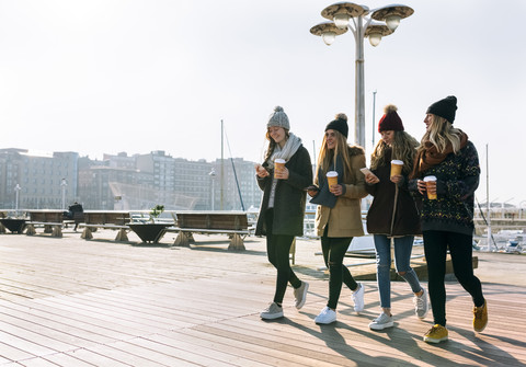 Vier Freunde mit Kaffee zum Mitnehmen und Handys beim Spaziergang auf der Promenade im Winter, lizenzfreies Stockfoto