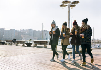 Vier Freunde mit Kaffee zum Mitnehmen und Handys beim Spaziergang auf der Promenade im Winter - MGOF02948