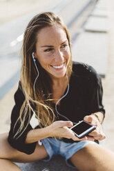 Happy young woman with cell phone and earbuds outdoors - GIOF01822