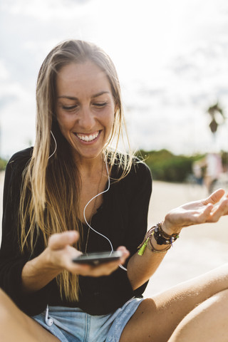 Glückliche junge Frau mit Handy und Ohrstöpseln im Freien, lizenzfreies Stockfoto