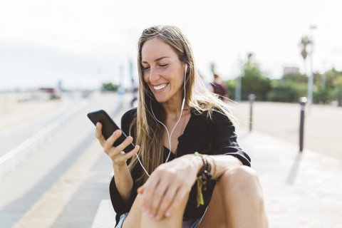 Lächelnde junge Frau mit Handy und Kopfhörern an der Uferpromenade, lizenzfreies Stockfoto