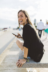 Happy young woman with cell phone and earbuds on waterfront promenade - GIOF01825