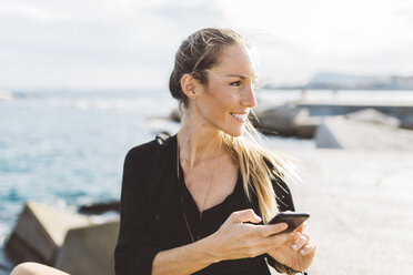 Lächelnde junge Frau mit Handy an der Strandpromenade, die sich umschaut - GIOF01835