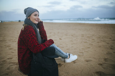 Lächelnde junge Frau, die im Winter am Strand sitzt - KIJF01188