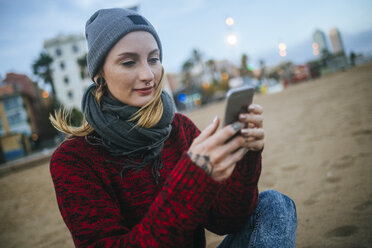 Junge Frau sitzt im Winter am Strand und benutzt ein Mobiltelefon - KIJF01189