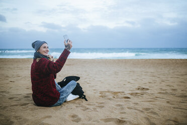 Lächelnde junge Frau, die im Winter am Strand sitzt und ein Mobiltelefon benutzt - KIJF01192