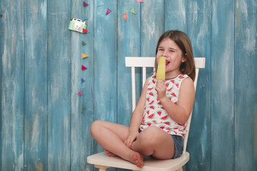 Glückliches kleines Mädchen sitzt auf einem Stuhl und isst einen Zitroneneislutscher - RTBF00649