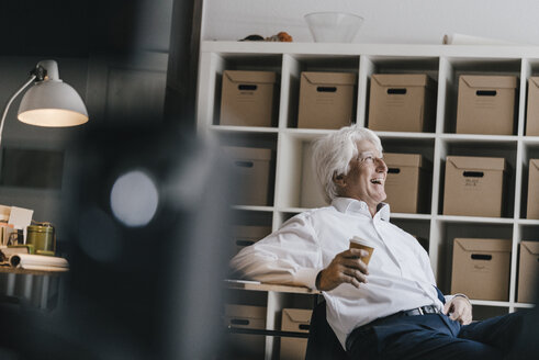 Lachender älterer Geschäftsmann mit Kaffee zum Mitnehmen in seinem Büro sitzend - KNSF01001