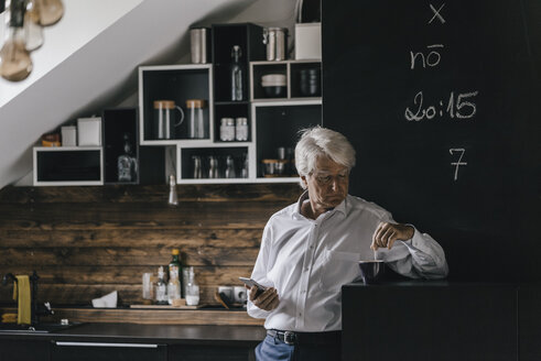 Älterer Geschäftsmann mit Smartphone und einer Tasse Kaffee in der Küche - KNSF01003