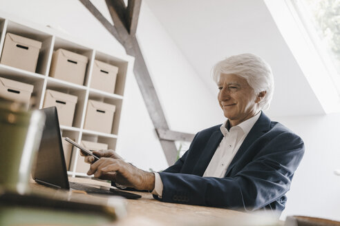 Smiling senior businessman using laptop in his office - KNSF01014