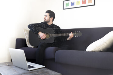 Junger Mann sitzt zu Hause auf der Couch und spielt Gitarre - FMOF00148