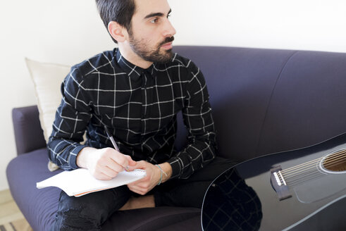 Junger Mann zu Hause auf der Couch sitzend mit Notizblock und Gitarre - FMOF00152