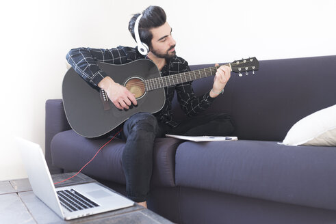 Junger Mann zu Hause, der Gitarre spielt und Kopfhörer trägt, die an einen Laptop angeschlossen sind - FMOF00153