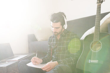 Young man at home with guitar wearing headphones and taking notes - FMOF00160