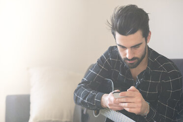 Junger Mann zu Hause mit Gitarre und Mobiltelefon - FMOF00172