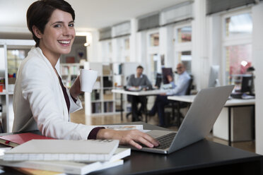 Porträt einer glücklichen Frau, die einen Laptop im Büro benutzt, mit Kollegen im Hintergrund - FKF02134