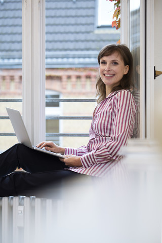 Porträt einer lächelnden Frau mit Laptop am Fenster, lizenzfreies Stockfoto