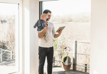 Father standing at home, carrying baby son and reading text messages - UUF09871