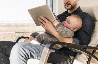 Vater im Sessel sitzend mit Baby auf dem Schoß, mit digitalem Tablet - UUF09894