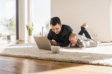 Vater mit kleinem Sohn benutzt Laptop auf Teppich liegend - UUF09903