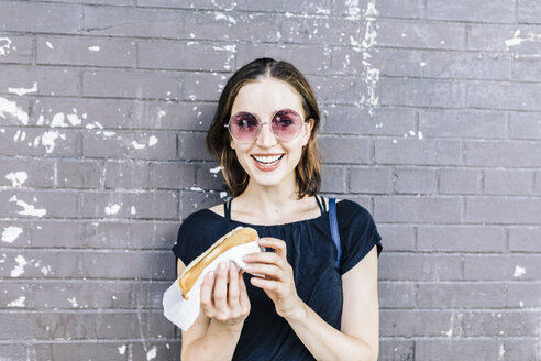 Porträt einer lächelnden Frau mit Hot Dog vor einer Wand - GIOF01866