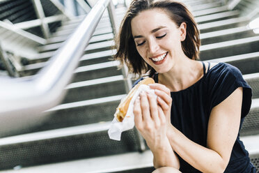 Lächelnde Frau mit Hot Dog auf einer Treppe sitzend - GIOF01871
