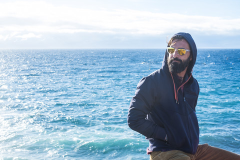 Porträt eines Mannes mit verspiegelter Sonnenbrille und Kapuzenjacke vor dem Meer, lizenzfreies Stockfoto