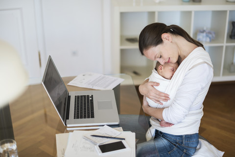 Mutter mit Baby im Tragetuch arbeitet von zu Hause aus, lizenzfreies Stockfoto