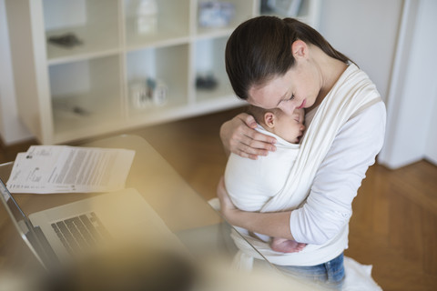 Mutter mit Baby im Tragetuch arbeitet von zu Hause aus, lizenzfreies Stockfoto