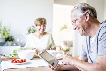 Älterer Mann zu Hause, der ein digitales Tablet benutzt, während eine Frau im Hintergrund einen Salat zubereitet - WESTF22756