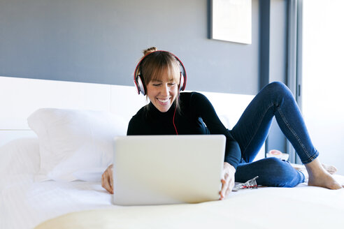 Lächelnde junge Frau, die auf dem Bett liegt, einen Laptop benutzt und Kopfhörer trägt - VABF01111
