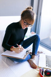 Smiling young woman on bed writing on clipboard - VABF01117