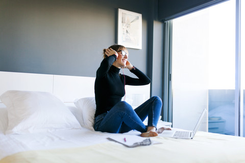 Glückliche junge Frau auf dem Bett mit Laptop und Kopfhörern, lizenzfreies Stockfoto