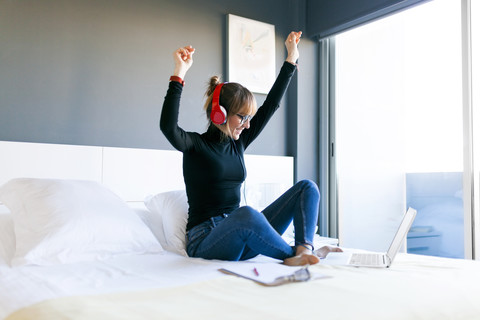 Glückliche junge Frau auf dem Bett mit Laptop und Kopfhörern, lizenzfreies Stockfoto