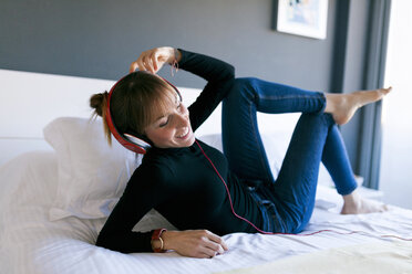 Happy young woman lying on bed wearing headphones - VABF01124