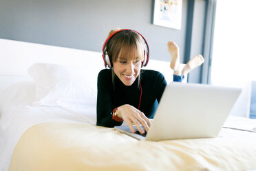 Lächelnde junge Frau, die auf dem Bett liegt, einen Laptop benutzt und Kopfhörer trägt - VABF01125