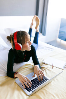 Young woman lying on bed using laptop and wearing headphones - VABF01126