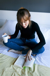 Junge Frau sitzt auf dem Bett, trinkt eine Tasse Kaffee und liest ein Buch - VABF01134