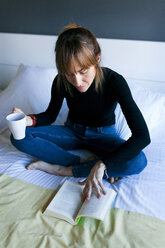 Young woman sitting on bed drinking cup of coffee and reading a book - VABF01134